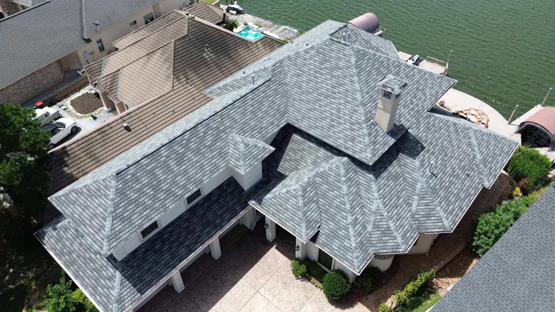 Aerial drone view of a luxury lakefront home with a gray shingle roof, pool, and private boat dock